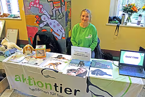 Eröffnung des Tierschutzzimmers in der Zinnowwald Grundschule in Berlin Zehlendorf Eine Frau im grünen Pullover sitzt an einem Tisch mit aktion tier-Banner und Informationsmaterialien darauf.