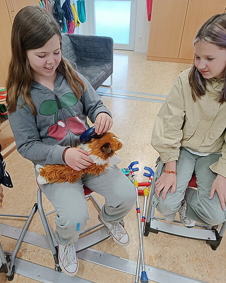 Zwei Mädchen auf einem Stuhl. Eines kämmt einen Stoffhund.