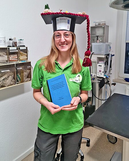 Eine lächelnde Frau mit Doktorhut steht mit ihrer Doktorarbeit in den Händen in einem Behandlungsraum.