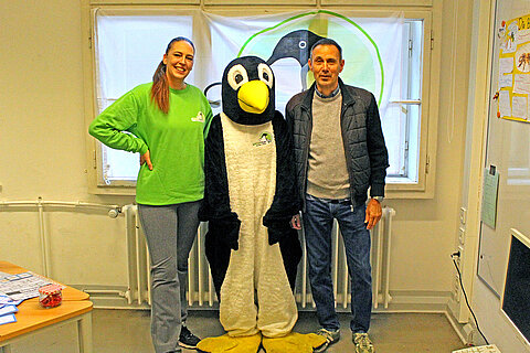 Eröffnung des Tierschutzzimmers in der Zinnowwald Grundschule in Berlin Zehlendorf Eine Frau in grünem Pullover, ein Pinguin-Maskottchen und ein Mann stehen vor einem Fenster.